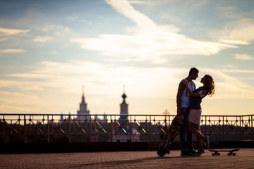 Outdoor shot of affectionate young couple pose against beautiful city view and sky, have rest after skateboading, demonstrate true love and perfect relationships. Talented skateboarders togehter.