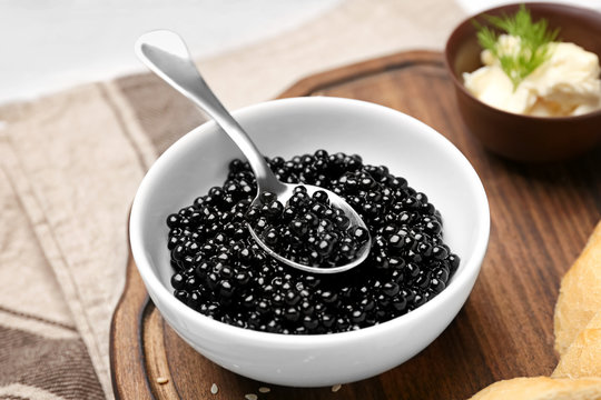 Bowl and spoon with delicious black caviar on wooden board, closeup