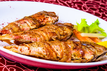 Grilled surmullet fish wrapped in bacon on white plate. Close-up