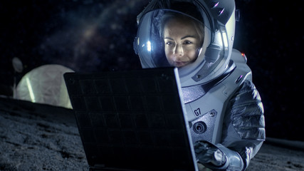 Female Astronaut Wearing Space Suit Works on a Laptop, Exploring Newly Discovered Planet, Communicating with the Earth. In the Background Space Habitat. Colonization Concept.