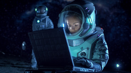 Obraz na płótnie Canvas Female Astronaut Wearing Space Suit Works on a Laptop, Exploring Newly Discovered Planet, Communicating with the Earth. In the Background Her Crew Member and Living Station. Colonization Concept.