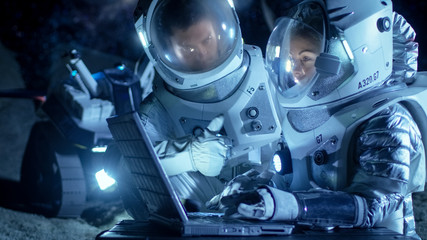 Two Astronauts Wearing Space Suits Work on a Laptop, Exploring Newly Discovered Planet,...
