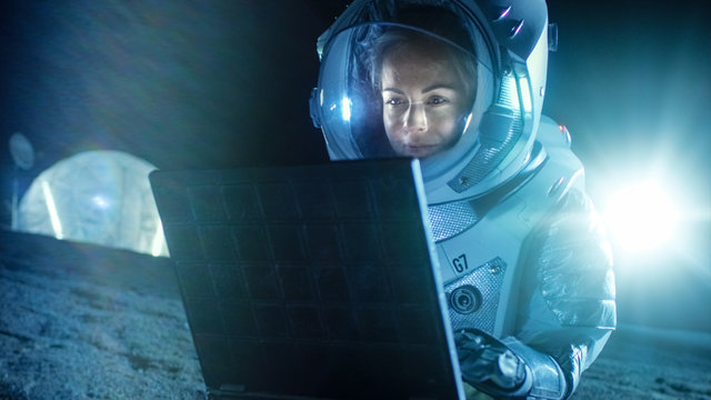 Female Astronaut Wearing Space Suit Works on a Laptop, Exploring Newly Discovered Planet, Communicating with the Earth. In the Background Space Habitat. Colonization Concept.