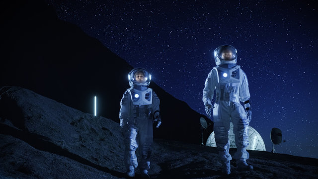 Two Astronauts in Space Suits Exploring Newly Discovered Planet. In the Background Space Base. Night Sky Full of Stars. Concept on Space Colonization.