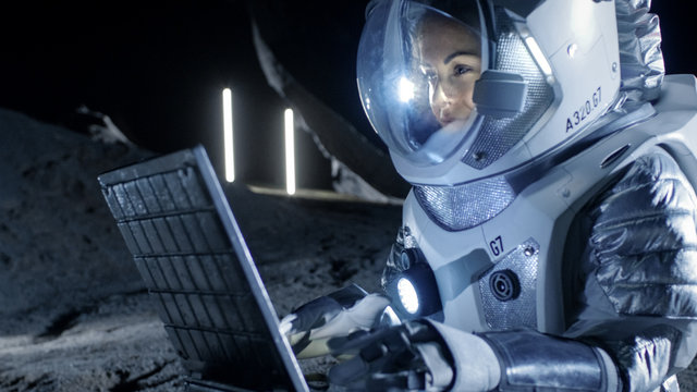 Female Astronaut Wearing Space Suit Works on a Laptop, Exploring Newly Discovered Planet, Communicating with the Earth. In the Background Her Space Habitat. Colonization Concept.