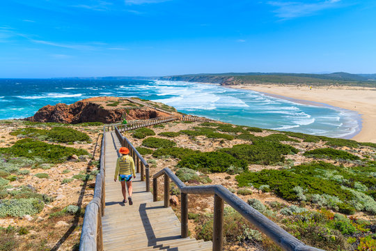 Young woman tourist waking on wooden walkway to beautiful Praia da Bordeira beach, popular place to do kite surfing, Algarve, Portuga