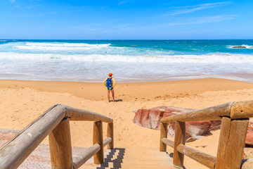 Walkway to sandy Praia do Amado beach and young woman tourist standing on sea shore, Portugal