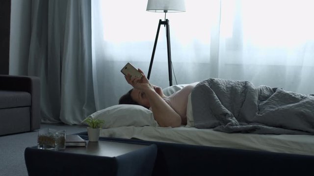 Charming middle aged long brown hair woman in pajams waking up with mobile alarm clock and stretching her arms in the bed in the morning. Yawning plus size female stretching in the bed after awake.