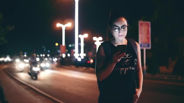 Attractive woman using a mobile phone while walking through the streets in a night city, in the background can see bikers. Stock footage.