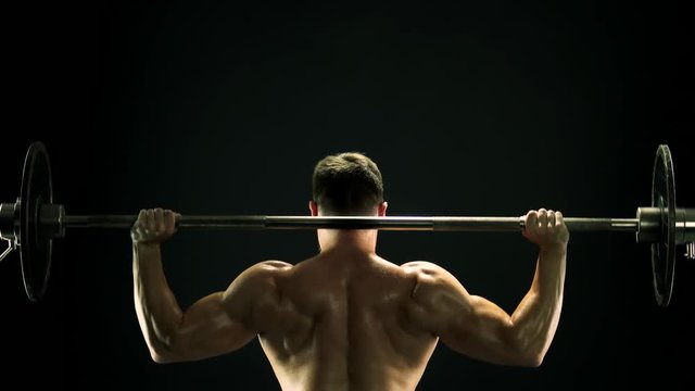Muscular man lifting barbell, back view. Rear view sweaty muscular sportsman lifting heavy weight on dark background. Professional bodybuilding concept.