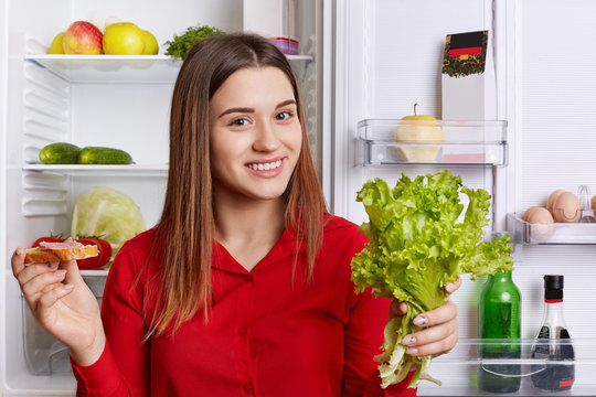 Attractive woman wears fashionable red blouse, stands near opened fridge, holds lettuce and sandwhich, going to prepare delicious dinner. Female cooker poses near refrigerator in kitchen at home