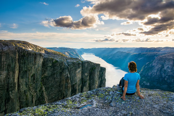 A woman is sitting on the edge of cliff on the way to boulder (Kjeragbolten) stuck in between the mountain crevices of Kjerag above a fjord, near Lysebotn, Norway. The feeling of complete freedom