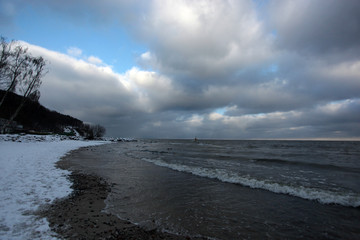 Baltic sea winter landscape - water waves, sandy and  snowy beach, with black big rock in the background with bright blue  sky with clouds on a sunny winter day
