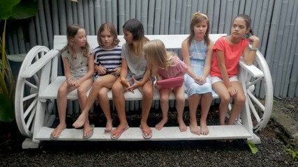 Two cliques of young girls one happy one bored on a long bench