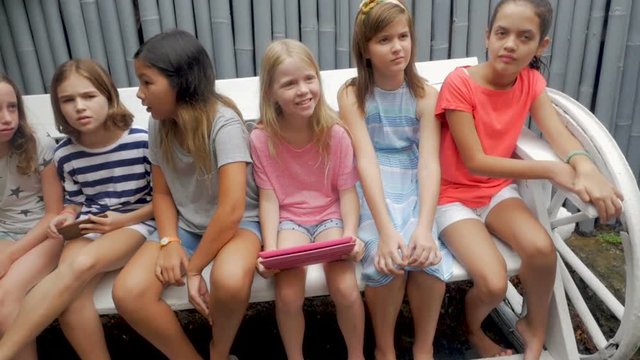 Six young pre teenage girls sitting on a bench holding digital technology