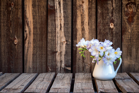 A bunch of freesia flowers in a white ceramic pitcher on a rustic wooden plank table.
