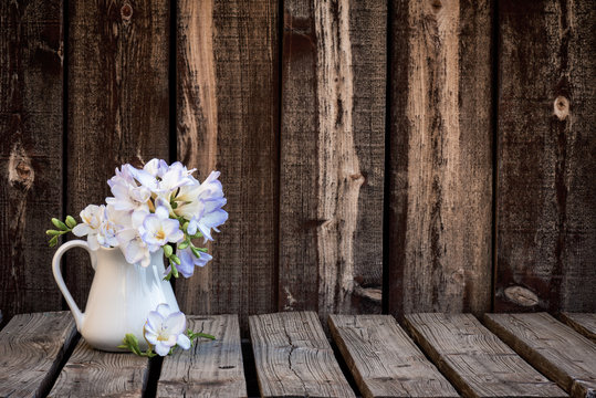 A bunch of freesia flowers in a white ceramic pitcher on a rustic wooden plank table.