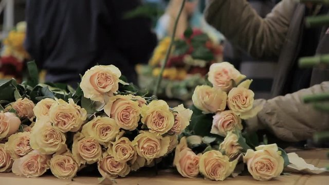 Man is arranging roses into a bunch on streen market. Male florist is standing at the counter covered with brown cardboard and taking egg shell flovers with long green scapes form it by his hands in