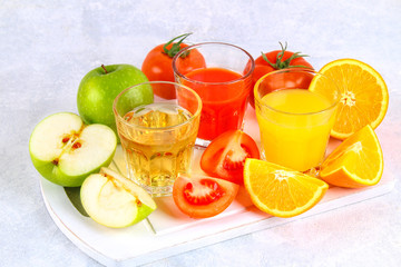 Glasses with fresh orange, apple, tomato juice on a gray concrete table. Lobules Fruits and vegetables around.