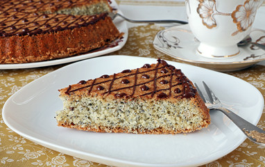  A piece of sliced pie with nuts and poppy seeds decorated with chocolate icing,on a white plate. Close Up, on the background of a pie and a saucer with a cup.