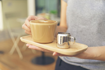 Young barista woman holding wooden brown tray with hot coffee and syrup in stainless cup and serving to customer in morning at coffee shop.
