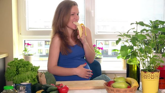 Beautiful caucasian pregnant woman eating banana in kitchen