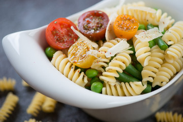 Closeup of fusilli with fresh cherry tomatoes, green peas, beans and parmesan served in a white bowl, selective focus, shallow depth of field