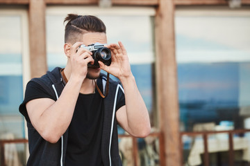 In search of best angle to create masterpiece. Portrait of dreamy good-looking caucasian guy in trendy outfit taking photos on street, looking through camera, focusing on model to make great shot