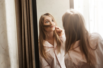 Fooling around while nobody see. Portrait of adorable playful blonde woman making moustache from hair strand, looking in mirror and making funny face, wearing cozy nightwear while in her room