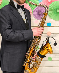 Naklejka premium saxophonist in an elegant grey suit, holds a saxophone in his hands and stands near a light wooden background