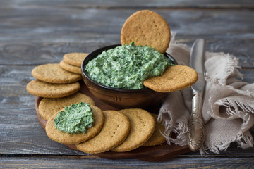 Green spinach dip with cream cheese, garlic and spices on a wooden board with oat crackers, rustic style, selective focus. Healthy homemade food