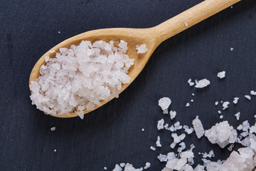 Crystal sea salt in a wooden spoon on dark background, top view, close-up, selective focus.