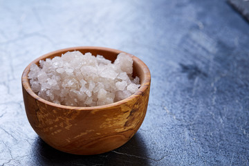 Large white sea salt in a natural wooden bowl on dark background, top view, close-up, selective...