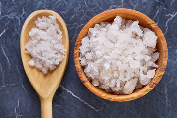 Large white sea salt in a natural wooden bowl on dark background, top view, close-up, selective focus