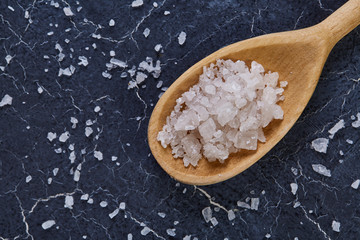 Crystal sea salt in a wooden spoon on dark marble background, top view, close-up, selective focus.