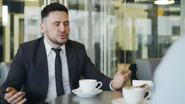 Angry bearded Caucasian businessman striking the table with his pen and scolding his employee for job mistakes in modern cafe.