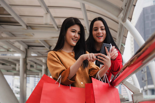 two beautiful cheerful asian and caucasian young woman with mobile smart phone, credit card and holding shopping bags together walking in modern city, sale, people, payment and shopping online concept