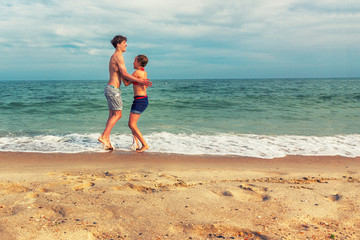 Fototapeta premium Two teenagers: a girl and a boy with blond hair, dressed in white T-shirts and shorts jumping up holding hands on a sea beach. Copy space.