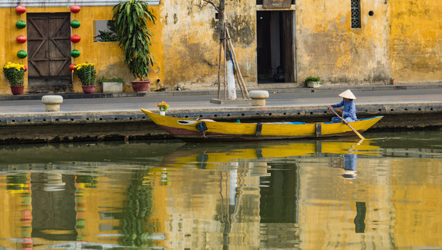 woman wearing iconic conical straw hat moving  her yellow wooden boat with oar  along river in old town of Hoi An, Vietnam