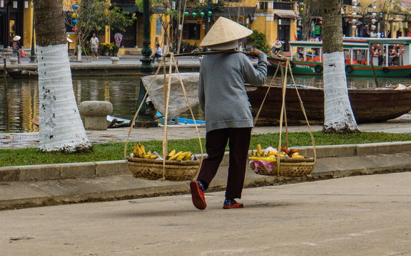 woman wearing iconic conical straw hat using a shoulder pole to carry baskets of fruit to sell