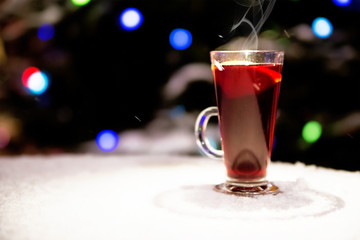 glass with mulled wine stands on the street on a snow table on the background of New Year's lights