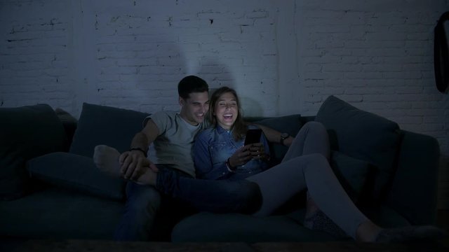 young happy and romantic couple on their 20s using together mobile phone enjoying sitting at home sofa couch browsing having fun with internet app in smart phone addiction concept