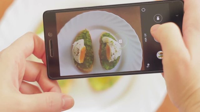 A woman in a restaurant takes a picture of food with a mobile phone camera. Sandwich with poached egg and avocado