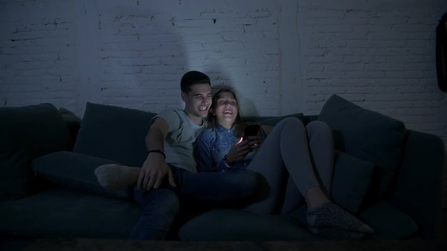 young happy and romantic couple on their 20s using together mobile phone enjoying sitting at home sofa couch browsing having fun with internet app in smart phone addiction concept