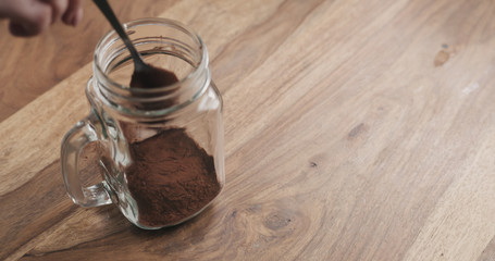 making cocoa drink in glass jar with handle on wood table, adding cacao powder