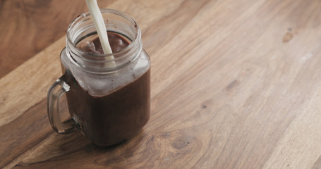 making cocoa drink in glass jar with handle on wood table, milk pour