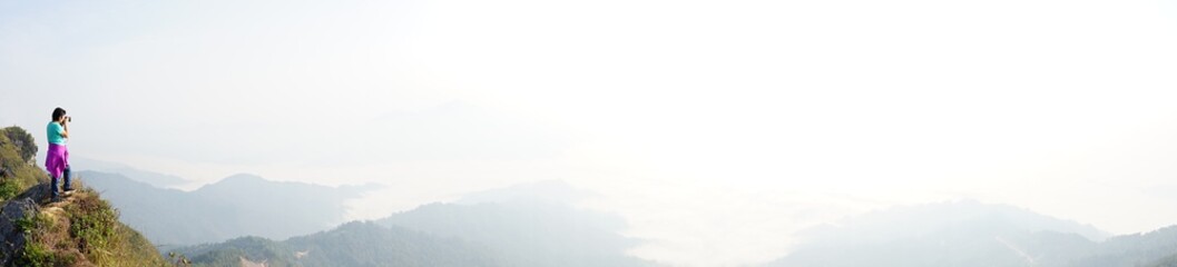 Panorama of woman holding camera and standing on the mountain top looking at the view of beautiful mountains with cloudy sky at  Doi Pha Tang, Chiangrai, Thailand