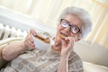 Elderly woman with plate of cookies eating delicious biscuit at home.