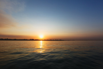 Beautiful sea landscape. Bright warm sunset in the sea near the coastline with the city's silhouettes. Sun hides behind the house. Colors of the sky are reflected in the water