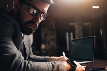 Close-up portrait of bearded man wearing eye glasses and smartwatch at night coworking loft space. Young creative manager looking away. Businessman using laptop and working at late evening